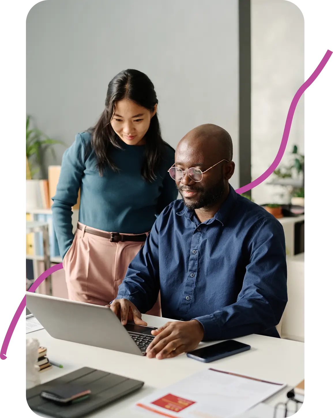 A man in glasses sits at a desk using a laptop, while a woman stands beside him, looking at the screen. They appear focused and collaborative.