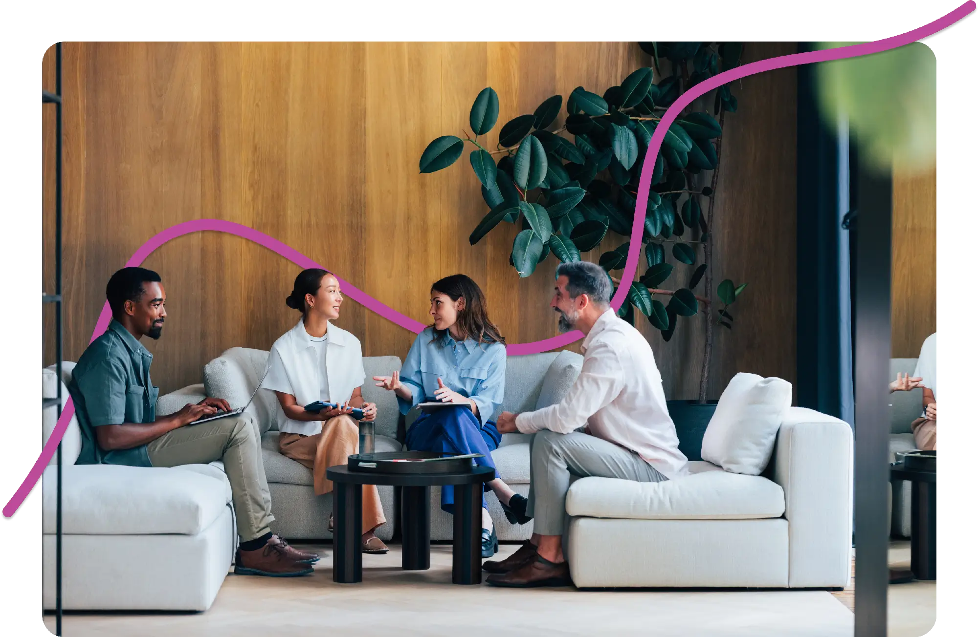 Four people sit on white sofas in a modern lounge, discussing and smiling. A large plant decorates the wooden wall. The atmosphere is relaxed and collaborative.