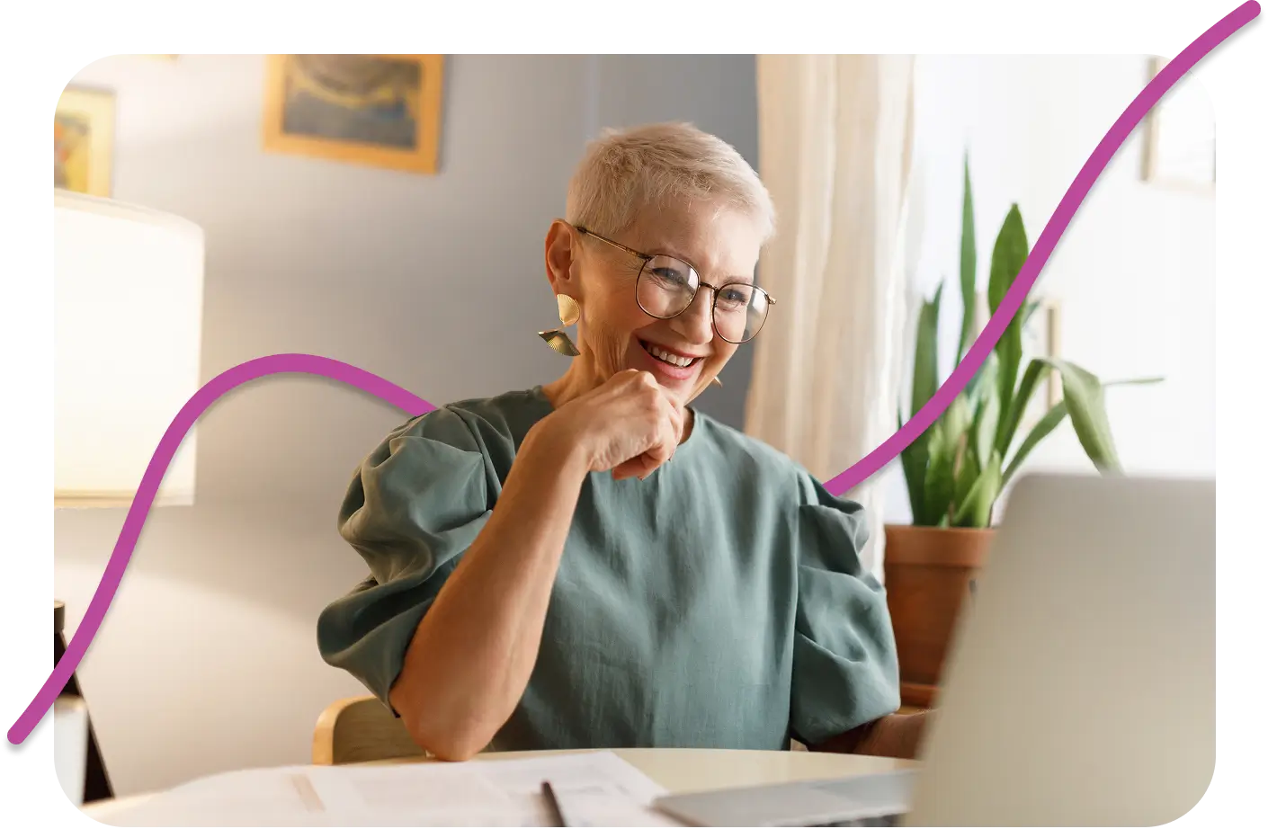A smiling elderly woman wearing glasses and a green blouse sits at a desk, looking at a laptop. A plant and lamp are in the background.