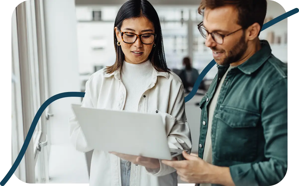 Two people with glasses, standing indoors, are discussing something on a laptop. They look engaged and focused.